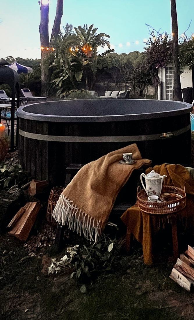 Hot tub de le&ntilde;a en el jard&iacute;n de Villa Paula al atardecer, con mantas y le&ntilde;a apilada junto a la estufa exterior